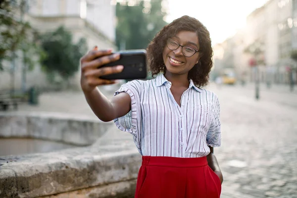 Portret van vrolijke jonge Afro-Amerikaanse zakenvrouw, in rode broek en gestreept shirt, selfie op haar smartphone buiten, poseren in de oude stad straat in de buurt van oude stenen fontein — Stockfoto