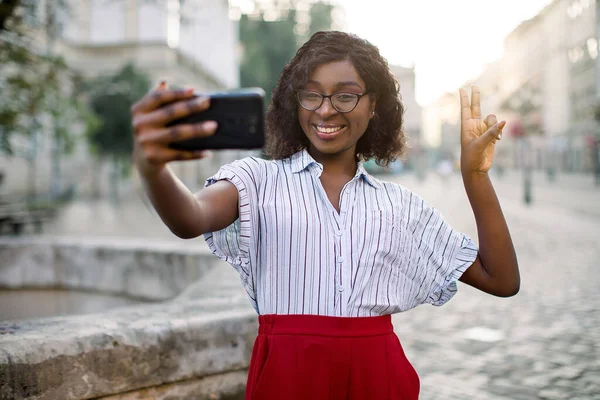 Outdoor stedelijke shot van mooie jonge afro-Amerikaanse glimlachende zakenvrouw, het maken van vrede teken en zelfportret op de smartphone, poseren op de oude stad straat. Selfie, mensen, lifestyle concept. — Stockfoto