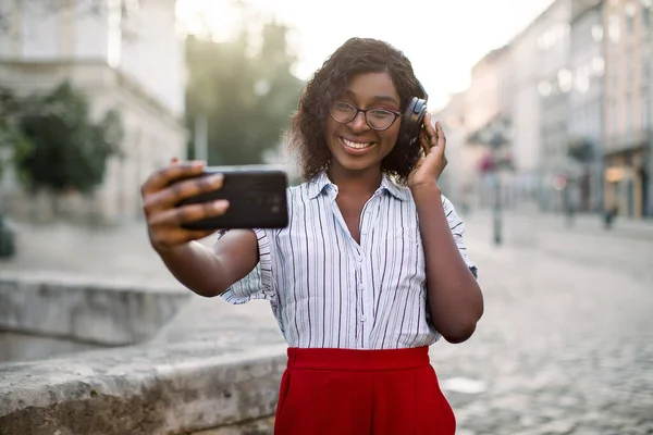 Lifestyle portret van jong gelukkig Afrikaans Amerikaans meisje met behulp van mobiele telefoon en koptelefoon, glimlachen, selfie foto 's maken en buiten muziek luisteren in de oude stad straat. Morgenstad — Stockfoto