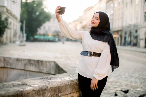 Side portrait of cheerful young Middle Eastern Muslim woman, wearing black hijab, pants and white shirt, taking selfie on her smartphone outdoors, posing in old city street near ancient stone fountain