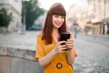 Sarı kıyafetler giyen, eski şehir caddesinde cep telefonuna mesaj yazan ve okuyan çekici beyaz bir kadının yatay portresini kapat. İnsanlar, teknoloji kavramı