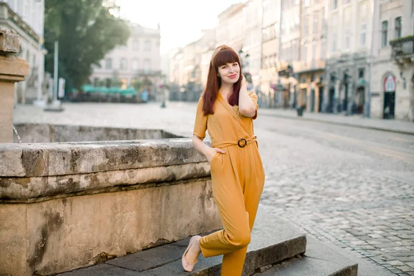 Affascinante giovane donna caucasica dai capelli scuri, indossa eleganti abiti gialli alla moda, sorridente alla macchina fotografica mentre in piedi su antica strada della città sulla vecchia fontana di pietra. Colpo città all'aperto. — Foto Stock