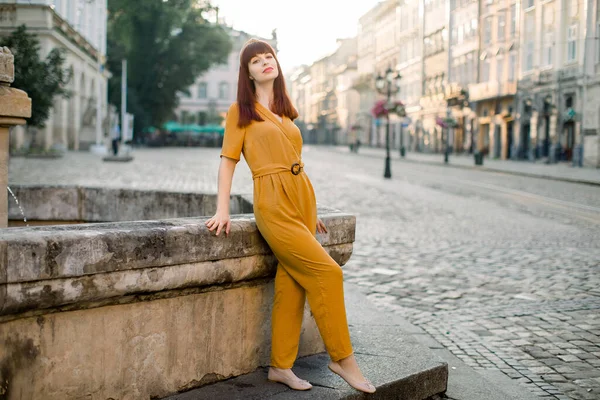 Scatto orizzontale di giovane bella donna caucasica in tuta gialla alla moda, in posa per la fotocamera con sorriso in ambiente urbano, in piedi vicino a antica fontana di pietra. Ritratto donna città — Foto Stock