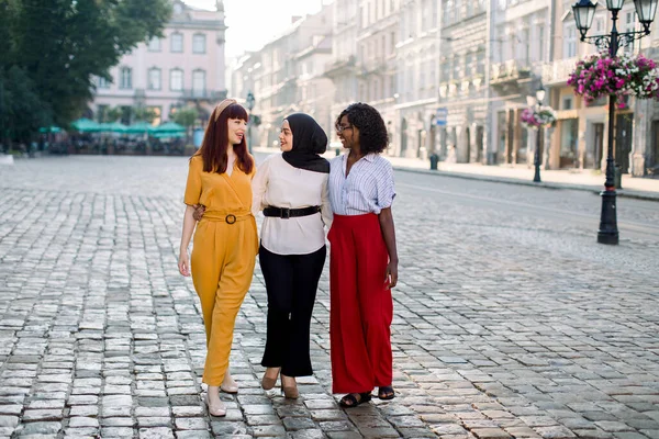 Outdoor full length portrait of three diverse multiethnical female friends walking down the ancient European city street. Three young women walking outdoors and having fun.