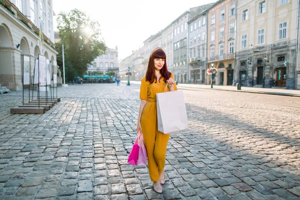 Retrato de moda de corpo inteiro ao ar livre de mulher elegante vestindo terno amarelo elegante e aro de cabeça, segurando sacos de compras coloridos enquanto caminhava na rua da cidade europeia — Fotografia de Stock