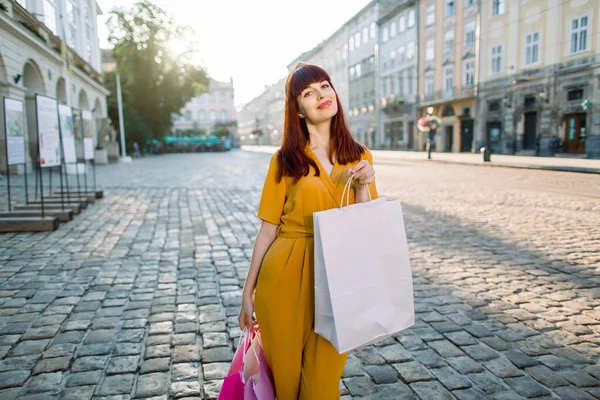 Outdoor urban portrait of young elegant fashionable woman wearing trendy yellow clothes, enjoying their walk and shopping in the city, smiling to camera and holding colorful shopping bags — Stock Photo, Image