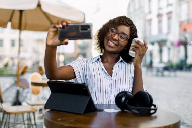 Çekici, çizgili gömlekli, genç Afrikalı esmer bir kadın. Selfie fotoğrafı çekiyor ya da video görüşmesi yapıyor. Şehir dışındaki kafede oturuyor, kahve içiyor ve dijital tablet kullanıyor.