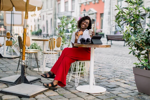 Mooie Afro-Amerikaanse vrouw, zitten in outdoor city cafe aan de tafel, met papier kopje koffie en croissant, genieten van haar lunchtijd en werk op tablet — Stockfoto