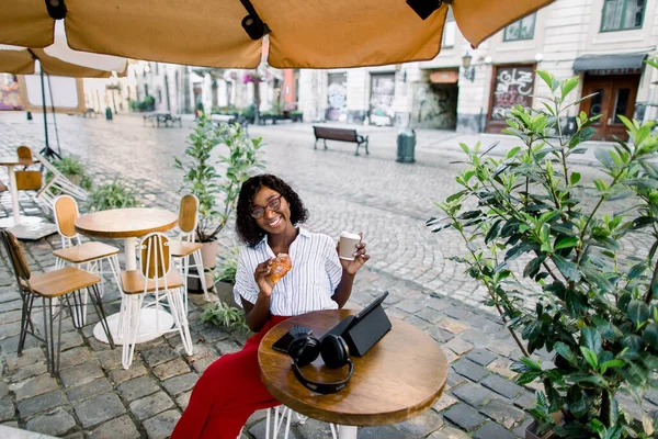 Top hoek portret van aantrekkelijke jonge Afrikaanse vrouw het drinken van koffie en het eten van verse croissant, zitten aan de tafel in outdoor city cafe, glimlachen om de camera — Stockfoto