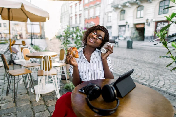 Mooie jonge Afro-Amerikaanse vrouw, zittend in outdoor city cafe, het vasthouden van papieren koffiebeker en verse smakelijke croissant, terwijl het werken op tablet. Zwarte dame buiten in de stad cafe — Stockfoto