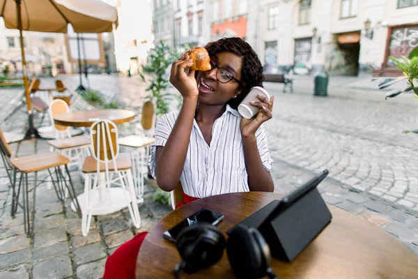 Portret van een grappige jonge Afrikaanse vrouw, die voor de camera speelt met afhaalkoffie en verse croissant, zittend in een koffiehuis buiten. Tablet ipad en koptelefoon op de tafel — Stockfoto