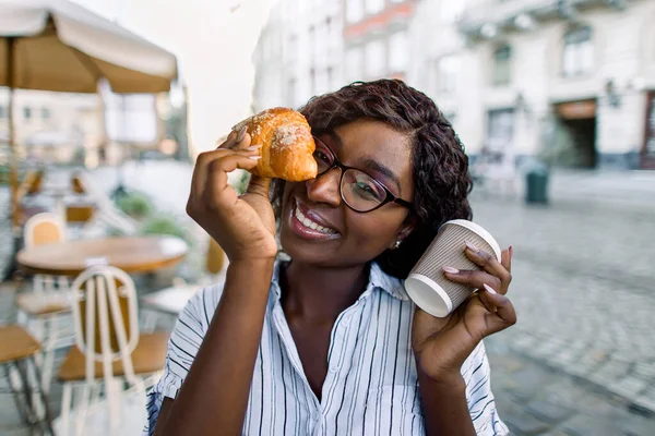 Outdoor stedelijke levensstijl portret van vrij schattig Afro-Amerikaanse dame, zittend in de coffeeshop met papier afhaalmaaltijd kopje koffie en verse croissant, glimlachen om de camera. — Stockfoto