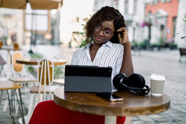 Attraente giovane intelligente concentrata donna africana è seduto in un ristorante all'aperto nella vecchia città europea, utilizzando tablet e cercando le informazioni in internet. Caffè da asporto, cuffie sul tavolo — Foto Stock