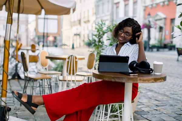 Portret van een gelukkige jonge Afrikaanse vrouw in slimme casual kleding, rode broek en gestreept shirt, ontspannen in een café en met behulp van een tablet. Lunch tijd in outdoor city cafe — Stockfoto