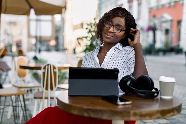 Portret van een mooie jonge Afrikaans-Amerikaanse vrouw in een bril, werkend met tablet ipad, zittend aan tafel in een koffiehuis, in de oude stad op zonnige dag — Stockfoto