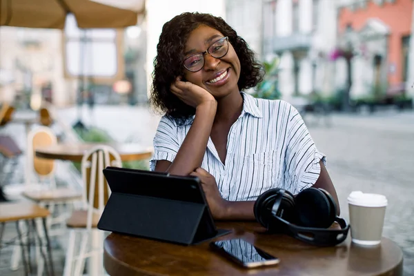 Mooie zwarte zakenvrouw, zittend aan de tafel in outdoor city coffee shop, met koptelefoon, smartphone en tablet ipad, genieten van haar lunchtijd buiten kantoor, glimlachen naar de camera — Stockfoto