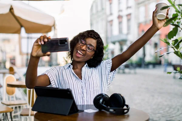 Portret van vrolijke lachende jonge Afrikaanse vrouw, zittend in een café in de oude Europese stad, videogesprek op mobiele telefoon, afhaalkoffie in de arm genomen. Tablet, koptelefoon op tafel. — Stockfoto