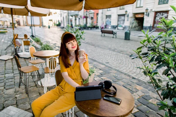 Ritratto angolo superiore di attraente giovane donna dai capelli rossi caucasici bere caffè da asporto, seduto al tavolo al caffè all'aperto della città, sorridente alla fotocamera, mentre si lavora su tablet — Foto Stock