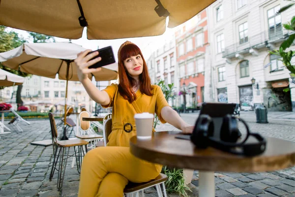 Charmante jonge mooie vrouw in gele overall, het maken van selfie foto of het hebben van video call, terwijl zitten in outdoor city cafe, het drinken van koffie en het gebruik van digitale tablet — Stockfoto