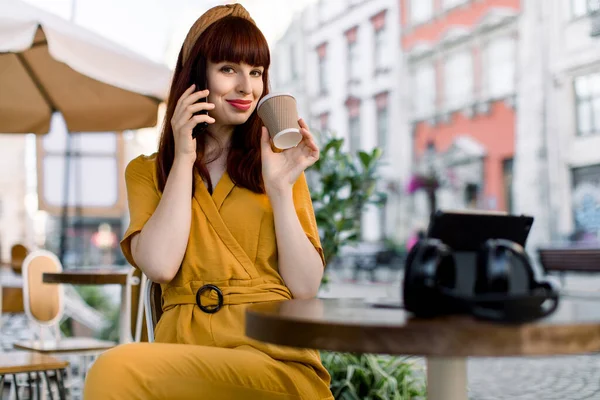 멋진 노란 수트를 입은 아름다운 빨간 머리 소녀가 시내 카페에서 스마트 폰으로 얘기하면서 카메라를 보고 커피를 마시고 있습니다. 뒤에 보이는 고대 건물들 — 스톡 사진