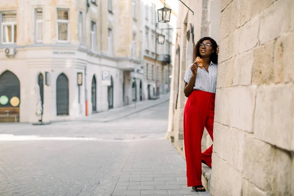 Encantadora jovem mulher africana em calças vermelhas, posando para câmera com croissant saboroso fresco, de pé ao ar livre no fundo de edifícios antigos da cidade vintage. — Fotografia de Stock