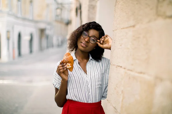 Menina afro-americana sorridente nova em óculos, segurando croissant saboroso, enquanto posando para câmera com sorriso, de pé perto de edifício da cidade de olf ao ar livre. Férias de verão, hora do almoço e conceito de férias. — Fotografia de Stock
