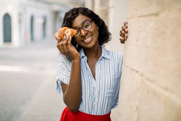 Close up urban lifestyle portrait of pretty cute African American lady, smiling to camera, funny hiding her eye with fresh croissant, standing outdoor on the background of old city buildings