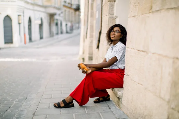 Horizontale shot van jonge aantrekkelijke dromerige Afrikaanse vrouw gekleed in rode broek en licht shirt, zittend op de trap van het oude vintage gebouw op straat buiten, met croissant — Stockfoto