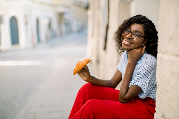 People, races, urban lifestyle and food concept. Close up portrait of cheerful smiling dark skinned African girl, wearing colorful casual clothes, sitting outdoor near the old building with croissant