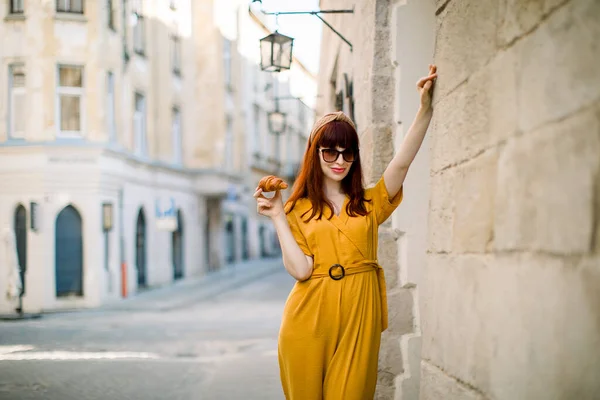 Portret miasta mody pięknej młodej białej, rudej kobiety, ubranej w stylowe, żółte ogrodniczki, okulary przeciwsłoneczne i obręcz głowy, spacerującej po starym europejskim mieście ze świeżym, smacznym rogalikiem — Zdjęcie stockowe