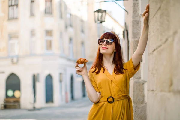 Ao ar livre retrato da cidade de sonho muito jovem caucasiana ruiva mulher, vestida com macacões amarelos elegantes, óculos de sol e aro cabeça, andando na velha cidade europeia com croissant saboroso fresco — Fotografia de Stock