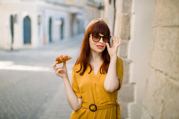 Horizontale Lifestyleaufnahme einer ziemlich glamourösen rothaarigen Frau, die stilvolle gelbe Kleidung, elegante Sonnenbrille trägt und mit frischem Croissant in der Hand auf der alten Stadtstraße spaziert. — Stockfoto