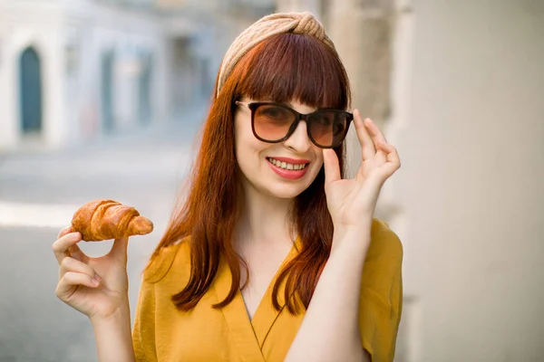 Nahaufnahme einer jungen attraktiven Frau in stylischer gelber Kleidung, Sonnenbrille und Reifrock auf dem Kopf, die ein Croissant in der Hand hält, ihren Stadtspaziergang genießt und in die Kamera lächelt. Verschwommene Altstadt im Hintergrund — Stockfoto
