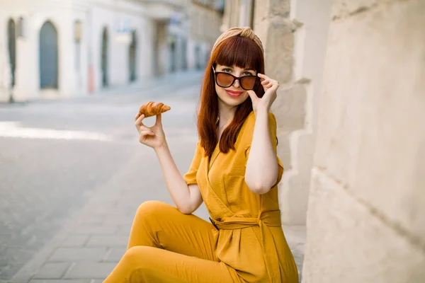 Streetfood, Tourismus, Mittagszeit und Urlaubskonzept. Junge lächelnde dunkelhaarige Kaukasierin in gelbem Anzug und Sonnenbrille, die Croissant isst, während sie auf der Treppe in der Stadt sitzt. Kopierraum — Stockfoto