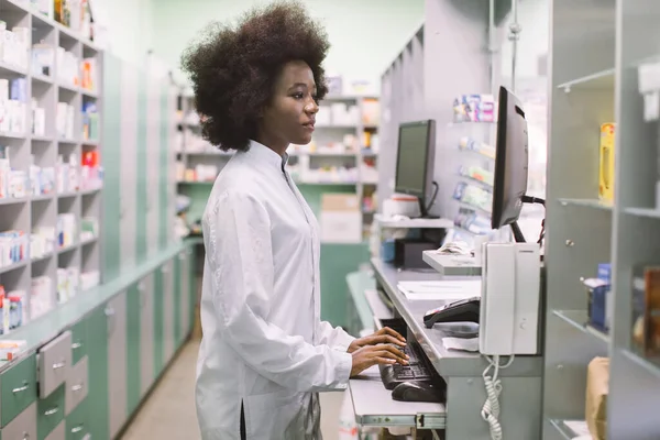 Boční pohled portrét mladé sebevědomé koncentrované africké lékárnice, práce s počítačem za pultem v lékárně — Stock fotografie