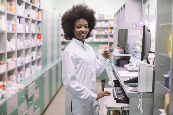 Afro-americká lékárnice na svém pracovišti v moderní lékárně, stojící poblíž pultu s počítačem a ukazující palec nahoru. Police s léky na pozadí — Stock fotografie