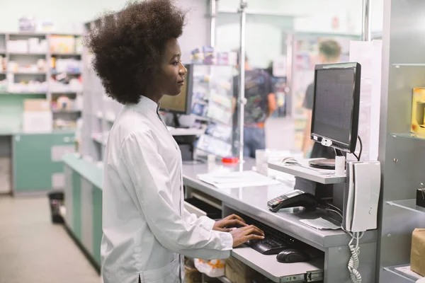 Usmívající se Afroameričanka klinický lékárník psaní na klávesnici počítače, zatímco stojí za pultem moderní nemocniční lékárny — Stock fotografie