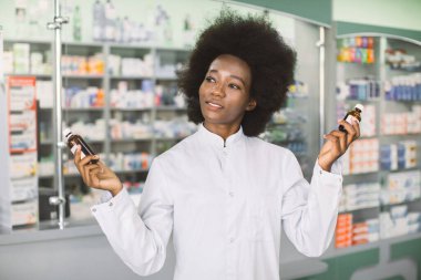 Eczane ve ilaç konsepti. Modern eczanede poz veren güzel Afrikalı Amerikalı kadın eczacının portresi, elinde iki şişe sıvı ilaçla