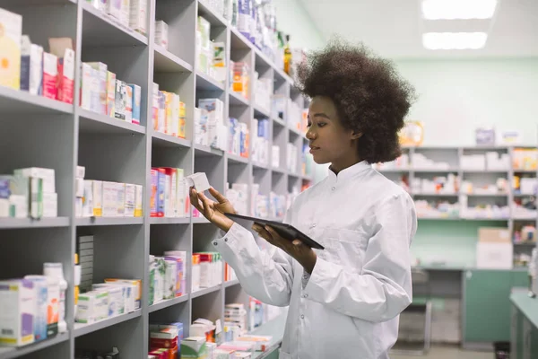 Mladá Afroameričanka lékárník nebo chemik pomocí digitální tablety, zatímco stojí v blízkosti polic s léky v moderní farmacii během zásob léků — Stock fotografie