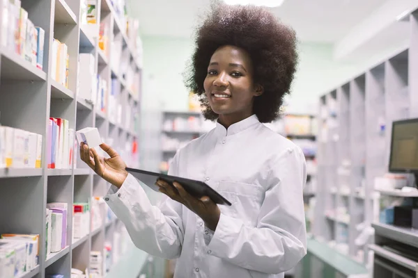 Afroamerykańska farmaceutka korzystająca z tabletu cyfrowego podczas inwentaryzacji w aptece, trzymająca pudełko z tabletkami i uśmiechnięta do kamery — Zdjęcie stockowe