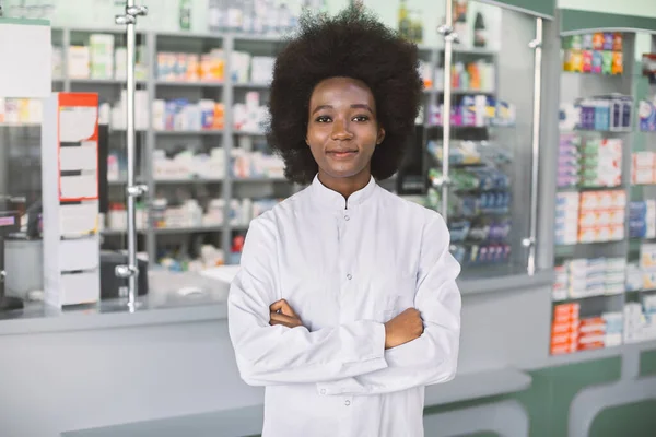 Pharmacy, healthcare concept. Young pretty smiling African dark skinned woman pharmacist in white coat, standing in interior of modern hospital pharmacy with arms crossed — Stock Photo, Image