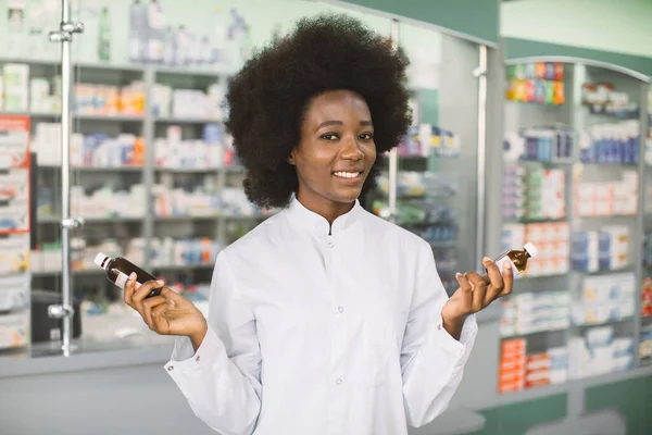 Pozitivní radostný mladý afroameričanka lékárník nebo lékař, držící v ruce dvě láhve s léky, stojící na pozadí moderní farmacie — Stock fotografie