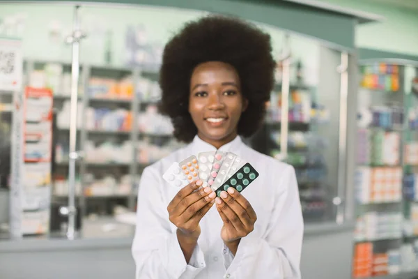 Joven farmacéutica afroamericana sonriente sosteniendo diferentes ampollas de medicamentos de colores en las manos, mientras está de pie en el interior de la farmacia moderna. Concéntrate en las manos —  Fotos de Stock