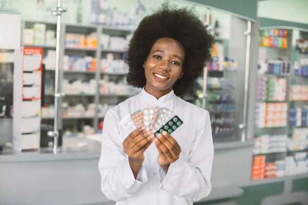 Mladá usmívající se Afroameričanka klinický lékárník drží různé barevné léky puchýře v rukou, zatímco stojí v interiéru moderní lékárny. Zaměření na obličej — Stock fotografie