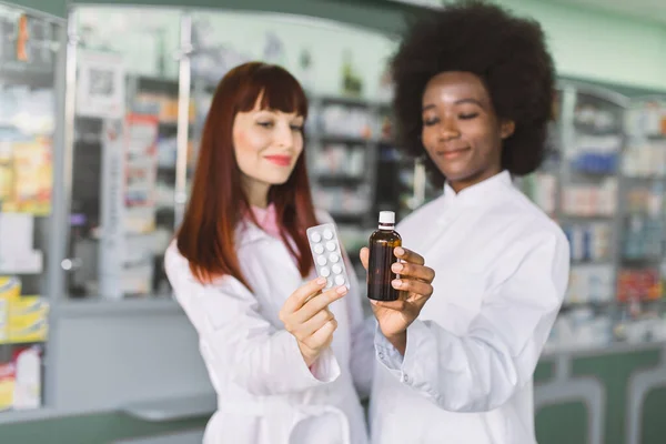 Dos farmacéuticas multiétnicas mujeres, africanas y caucásicas, que trabajan en farmacias, revisando la calidad de los medicamentos en la farmacia del hospital, mostrando a la cámara el frasco de jarabe y la ampolla de la píldora. Enfoque en drogas —  Fotos de Stock