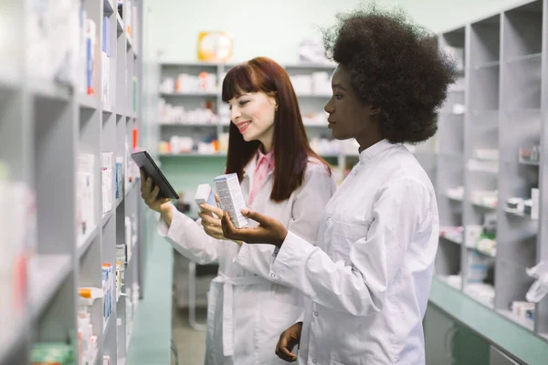 Medicine, pharmaceutics, health care and people concept. Two young multiethnic female pharmacist, African and Caucasian, taking medications from the shelf, using tablet in pharmacy — Stock Photo, Image