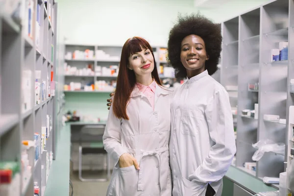 Two happy friendly multiethnic women clinical pharmacists or doctors, posing hugging each other together, smiling to camera in modern hospital pharmacy. Healthcare concept — Stock Photo, Image