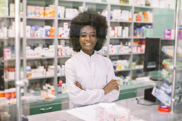 하얀 피부의 젊고 어두운 여자 약사 행복하게, 웃으면서 카메라 앞에 포즈를 취하고, 두 팔을 교차하여 약국 배경 위에 서 있습니다. 의약품, 약국, 사람들, 건강 관리 및 약리학 개념 — 스톡 사진