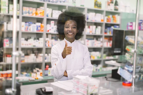 Retrato de una farmacéutica africana profesional sonriente en una farmacia moderna, parada detrás del vidrio en el mostrador, y mostrando su pulgar hacia arriba —  Fotos de Stock