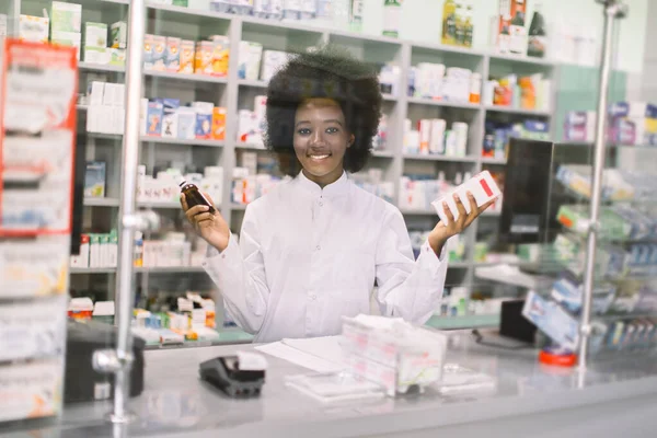 유리 뒤에 있는 젊고 젊은 아프리카 약사의 초상화, 카운터에 있는 현대 약국의 카메라 앞에서 웃고 있는 모습, 약을 손에 쥐고 있는 모습. 약과 의약품의 개념 — 스톡 사진